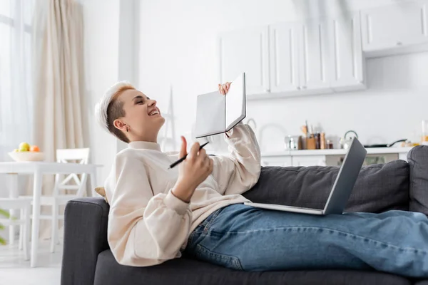 Excited woman with blank notebook laughing near laptop on couch — Stock Photo