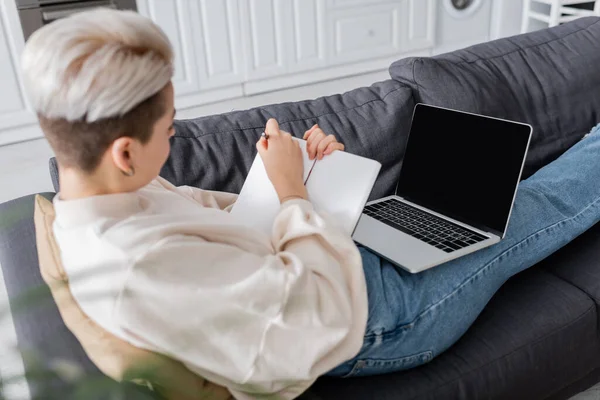 Blurred woman writing in notebook near laptop with blank screen on couch at home — Stock Photo