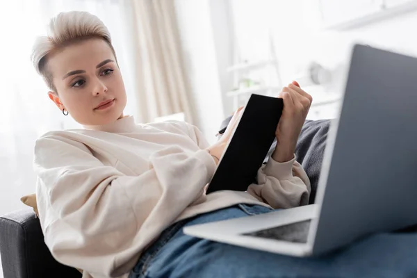 Mujer con la escritura de peinado de moda en el cuaderno en el sofá cerca de la computadora portátil borrosa — Stock Photo