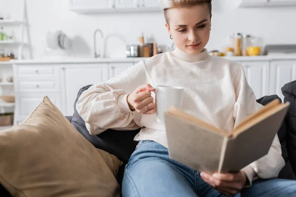 Giovane donna seduta sul divano con tazza di tè e libro di lettura — Foto stock