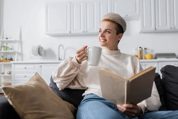 Mujer alegre con libro y taza de té sentado en el sofá en casa y mirando hacia otro lado - foto de stock