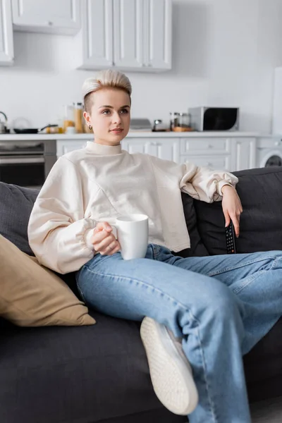 Trendy woman in white pullover holding cup of tea and watching movie on tv at home — стоковое фото