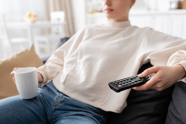 Partial view of blurred woman with tea cup and remote controller watching tv at home — Stock Photo