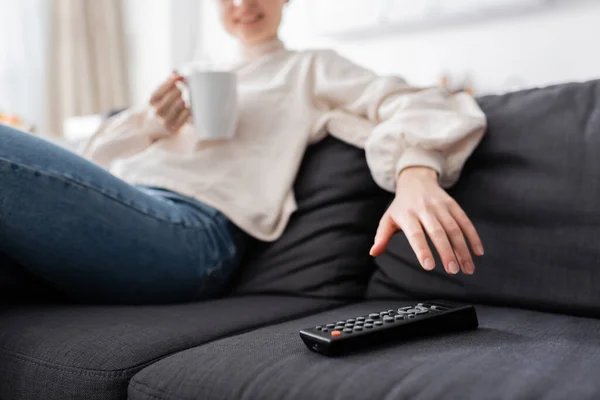 Vista parcial de la mujer borrosa con taza de té cerca del controlador remoto de televisión en el sofá - foto de stock