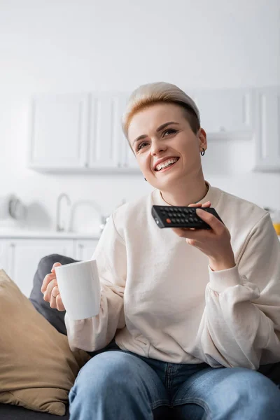 Sonriente mujer sosteniendo la taza de té mientras hace clic en los canales de televisión en casa - foto de stock