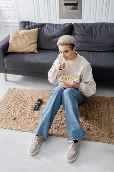 Vue grand angle de jeune femme manger du pop-corn tout en regardant la télévision sur le sol à la maison — Photo de stock
