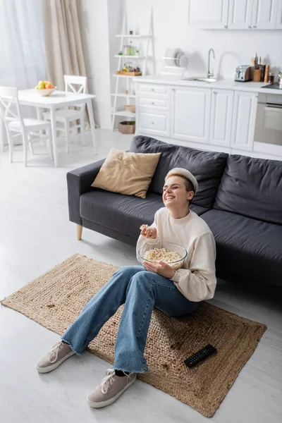 Donna allegra con ciotola di popcorn guardando film commedia sul pavimento in cucina a pianta aperta — Foto stock