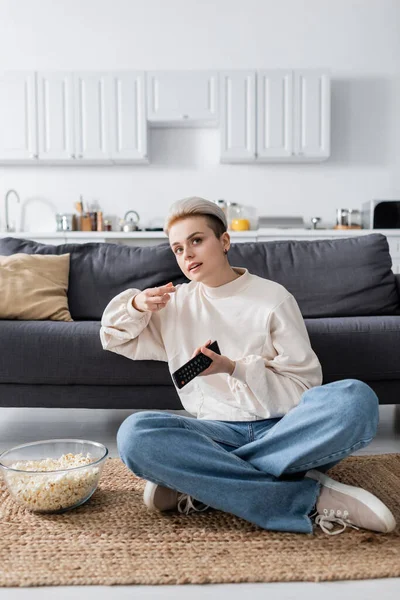 Mujer enfocada con control remoto de televisión comiendo palomitas de maíz y viendo películas en el suelo en casa - foto de stock