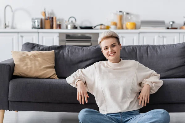 Cheerful woman in white pullover sitting near couch in open plan kitchen and looking at camera - foto de stock