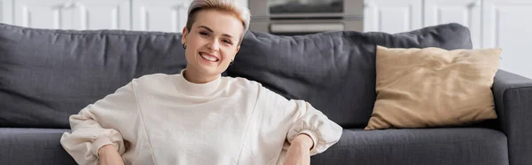 Happy woman in white pullover smiling at camera near couch and pillow, banner — Fotografia de Stock