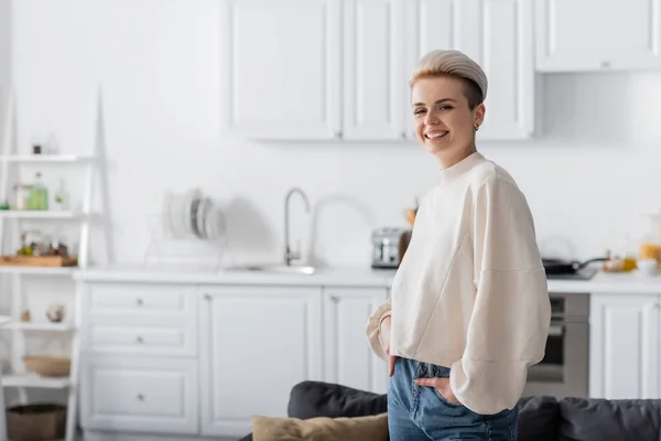 Mujer alegre en jersey blanco de pie en la cocina con las manos en los bolsillos y mirando a la cámara — Stock Photo
