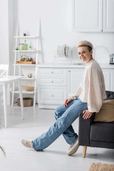 In voller Länge stilvolle Frau in Jeans sitzt in offener Küche und lächelt in die Kamera — Stockfoto