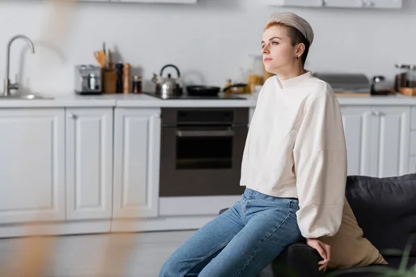 Dreamy woman in white pullover sitting in kitchen and looking away — стоковое фото