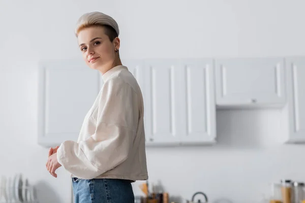 Donna positiva con acconciatura alla moda guardando la fotocamera in cucina sfocata — Foto stock