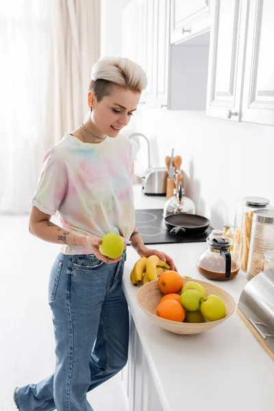 Trendy young woman holding apple and bananas near bowl with fresh fruits in kitchen — Stock Photo