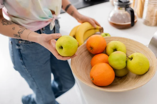 Cropped view of woman in jeans near bowl with fresh fruits and blurred coffee pot in kitchen — Stock Photo