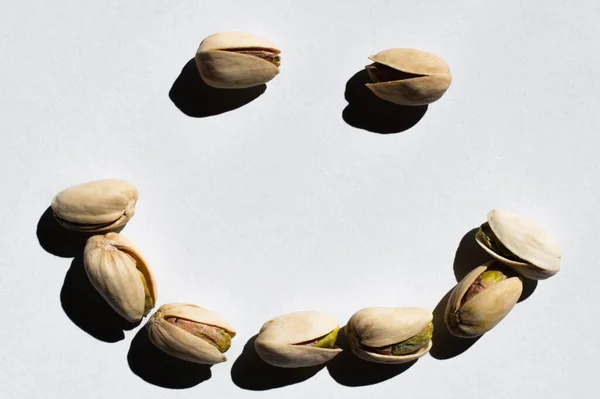 Flat lay of cracked green and salty pistachios in shape of smile on white background — стоковое фото