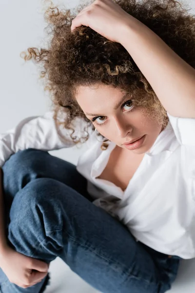 Vista ad alto angolo di bella donna riccia guardando la fotocamera e toccando i capelli su sfondo bianco — Foto stock