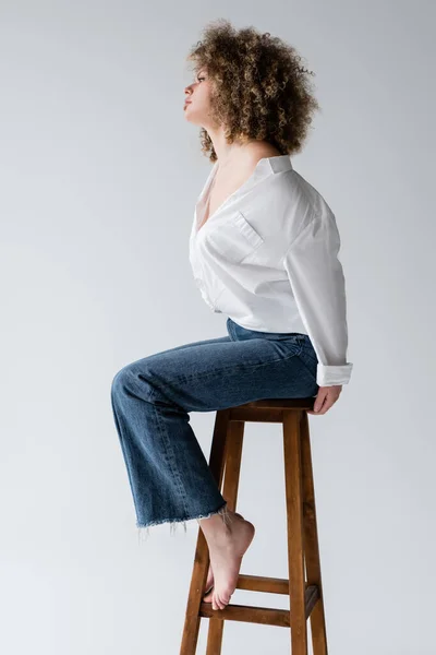 Vue latérale du modèle élégant pieds nus assis sur une chaise isolée sur blanc — Photo de stock