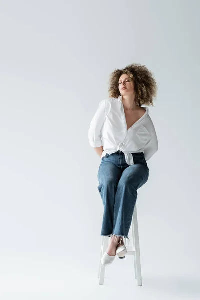 Modèle en chemisier et jeans assis sur une chaise sur fond blanc — Photo de stock