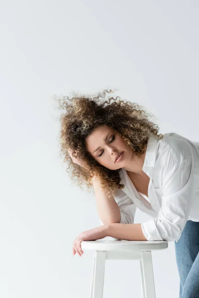 Jovem mulher tocando cabelo encaracolado perto da cadeira isolada no branco — Fotografia de Stock