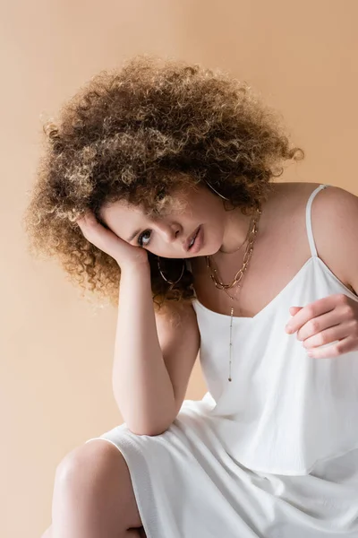 Modèle bouclé en vêtements blancs et colliers regardant la caméra isolée sur beige — Photo de stock