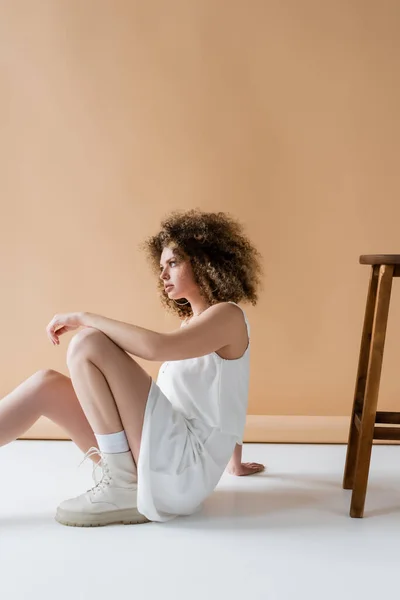 Trendy curly model in white clothes sitting near chair on beige background — Stock Photo