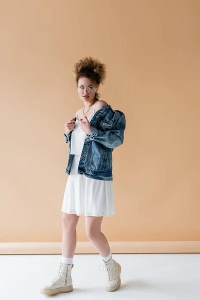 Trendy woman in eyeglasses and denim jacket on beige background — Stock Photo