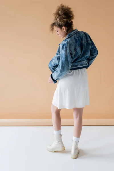 Vista laterale della donna riccia in giacca di jeans e stivali su sfondo beige — Foto stock