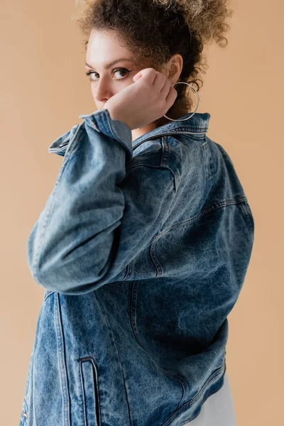 Кудрявая женщина в джинсовой куртке смотрит на камеру, изолированную от бежевого — стоковое фото