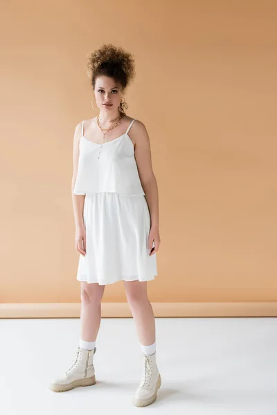 Полная длина стильная женщина в сапогах и белой одежде стоя на бежевом фоне — стоковое фото