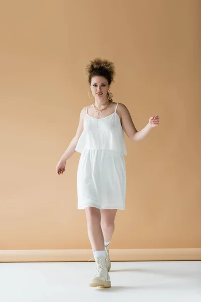 Mulher positiva em roupas brancas andando e olhando para a câmera no fundo bege — Fotografia de Stock