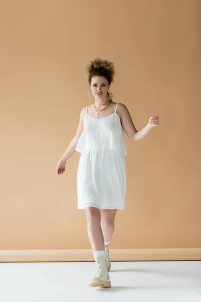 Positive woman in white clothes walking and looking at camera on beige background — Stock Photo