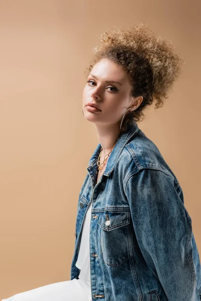 Curly model in denim jacket looking at camera isolated on beige — Stock Photo