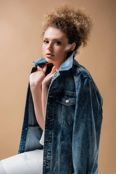 Молода жінка тримає джинсову куртку і дивиться на камеру ізольовано на бежевому — стокове фото