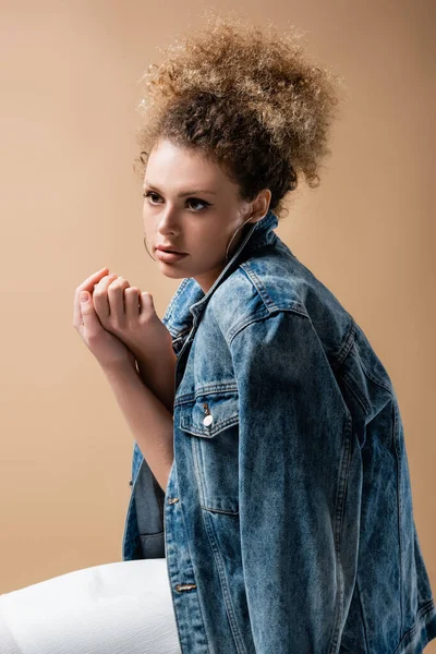 Stylish curly woman in denim jacket touching hands isolated on beige — Stock Photo