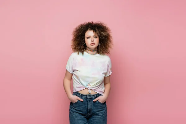 Young curly woman holding hands in pockets of jeans isolated on pink — Stock Photo