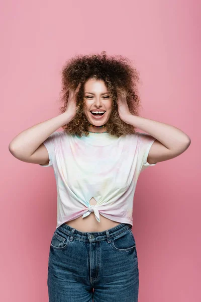 Heureuse femme bouclée touchant les cheveux isolés sur rose — Photo de stock