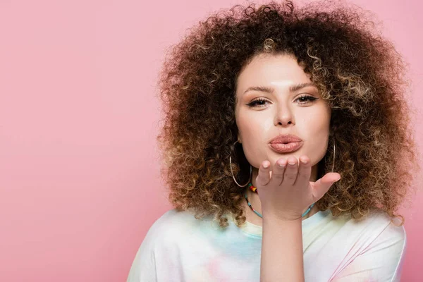 Portrait de jeune femme bouclée soufflant l'air baiser isolé sur rose — Photo de stock