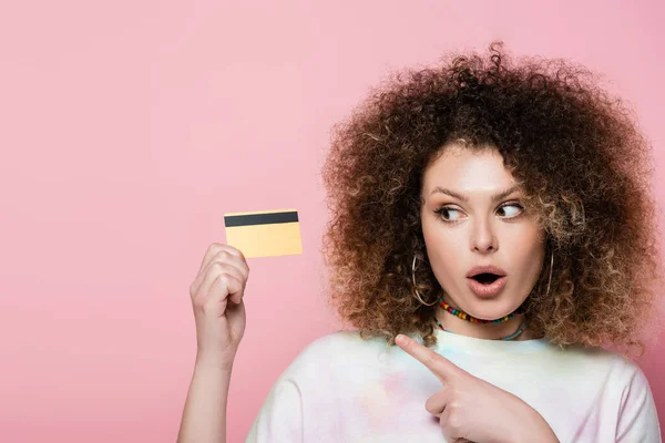 Mujer emocionada apuntando a la tarjeta de crédito con el dedo aislado en rosa - foto de stock