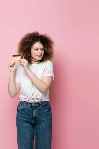 Mujer astuta sosteniendo la tarjeta de crédito y mirando hacia otro lado aislado en rosa - foto de stock