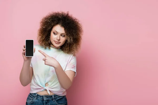 Bella e riccia donna che punta al cellulare isolato su rosa — Foto stock