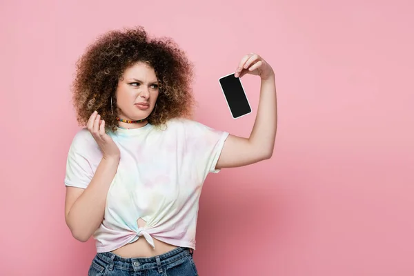 Mujer joven disgustada sosteniendo el teléfono móvil con pantalla en blanco sobre fondo rosa - foto de stock