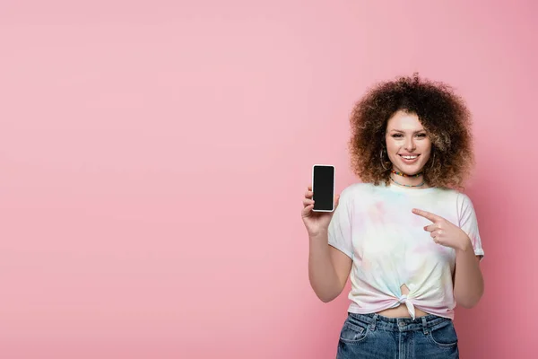 Mulher alegre apontando para o telefone celular isolado em rosa — Fotografia de Stock