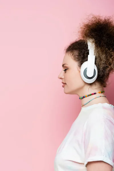 Seitenansicht einer lockigen Frau mit drahtlosen Kopfhörern auf rosa Hintergrund — Stockfoto