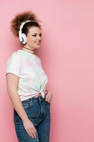 Mujer feliz en jeans y camiseta escuchando música en auriculares aislados en rosa - foto de stock