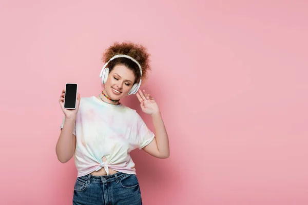 Curly woman in headphones holding cellphone on pink background — Stock Photo