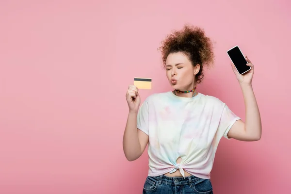 Кучерява жінка кидає губи, тримаючи кредитну картку і смартфон ізольовано на рожевому — стокове фото