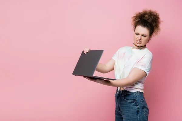 Disgusted curly woman holding laptop isolated on pink — Stock Photo