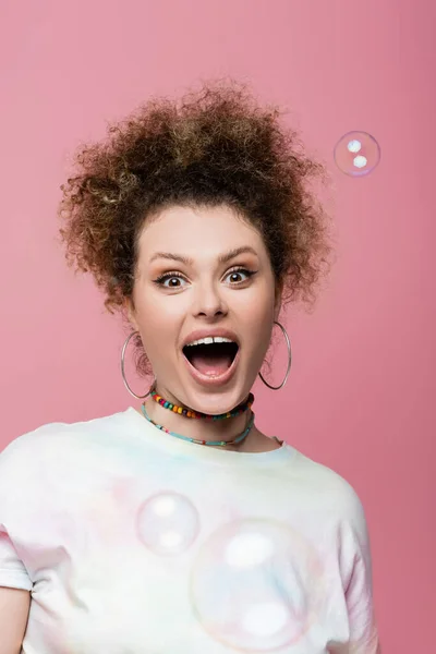 Excited curly woman looking at camera near soap bubbles on pink background — Stock Photo